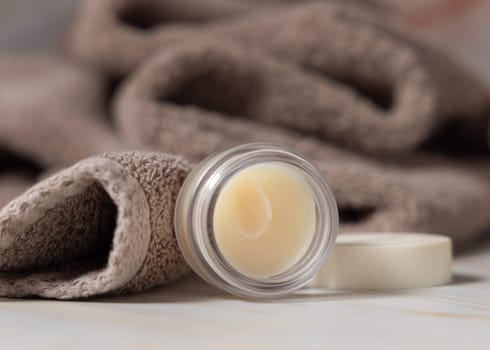 Opened lip balm jar with blank lid near brown bath towel closeup, cosmetic packaging mockup, copy space. Lipcare beauty product for mens and woman daily lipcare and personal care