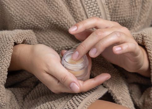 Close up of woman in brown bathrobe touching lip balmwith finger and holding opened cosmetic jar in other hand closeup, natural manicure. Organic cosmetics for body and face care