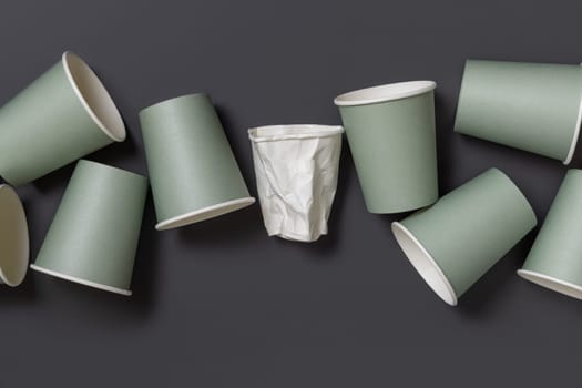 Disposable new light green paper cups and crumpled used white one in a line a dark gray top view, mockup with copy space. Empty cardboard coffee cups flat lay. No plastic and sustainable lifestyle concept