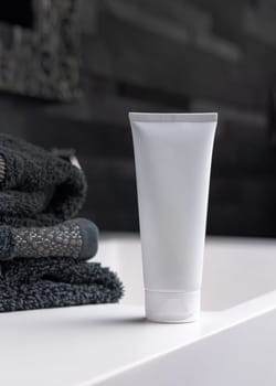 White cream squeeze tube near black folded towels on basin in dark grey masculine bathroom close up, packaging mockup. Natural plant unbranded cosmetics with copy space. Spa and wellness concept