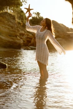 Beautiful young woman holding a starfish and enjoying it on the beach. The girl is standing in backlight. Her dress is glowing in the sun, in a white knitted dress with pearls on her neck