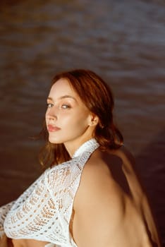 close-up beautiful slim young girl sits on sea beach, in sea. around her sea surface, she is dressed in white knitted dress, against background of big rocks. concept of summer sea morning