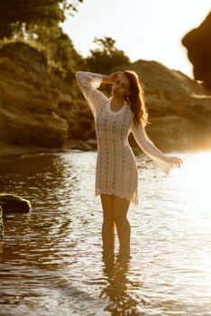 Beautiful young woman posing on the beach in summer. girl standing in backlight. her dress glows in the sun, in white knitted dress with pearls on her neck, in water against the backdrop of a rock