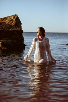 beautiful young girl posing on sea beach, in water. she is in sea, wearing white knitted dress, against background of big rocks. summer sea morning concept