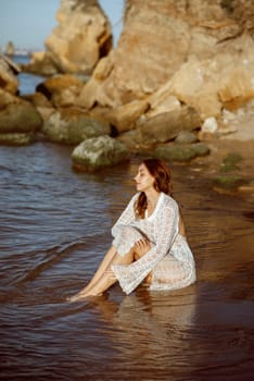 beautiful slim young girl sits on the sea beach, in the sea. around her the sea surface, she is dressed in a white knitted dress, against the background of large rocks. concept of a summer sea morning