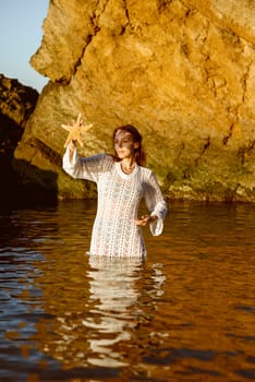 Beautiful young woman holding starfish in her hands and enjoying it on the beach. girl in white knitted dress with pearls on her neck. she stands in water against the backdrop of a rock