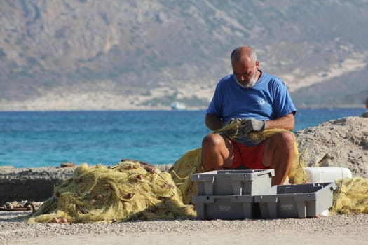 An adult man repairs tackle and fishing nets on the beach against a backdrop of mountains in Balos Bay, Greece