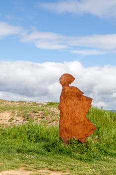 On the Camino de Santiago, metal sculptures of a pilgrim walking toward stunning hills under a cloudy sky.