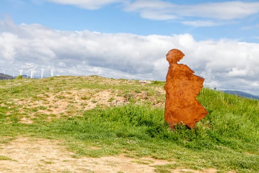 On the Camino de Santiago, metal sculptures of a pilgrim walking toward stunning hills under a cloudy sky.