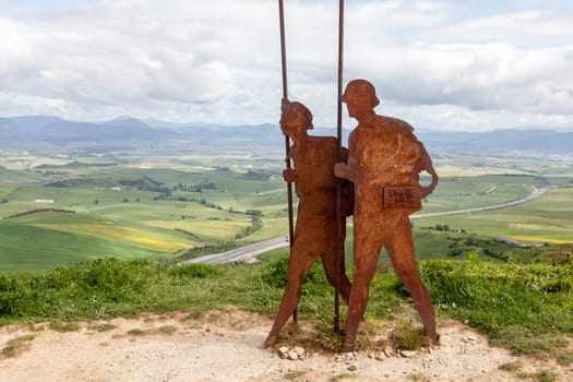On the Camino de Santiago, metal sculptures of pilgrims walk toward stunning hills under a cloudy sky.