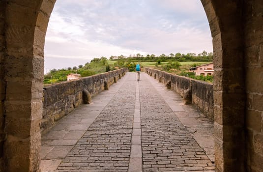 A person strolls on an ancient stone bridge in Obanos, Spain, enjoying the lush greenery and serenity of the landscape.
