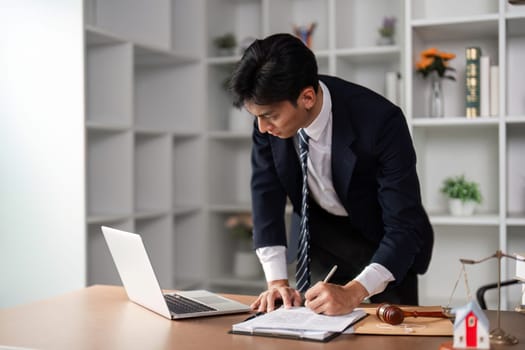 A young businessman is actively writing notes and reviewing documents while standing at his desk, demonstrating focus and professionalism in a contemporary office setting.
