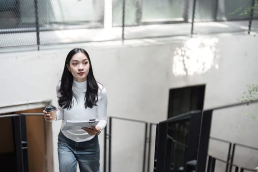 A young professional woman is ascending stairs while holding documents and a coffee cup, demonstrating focus and determination in her work.