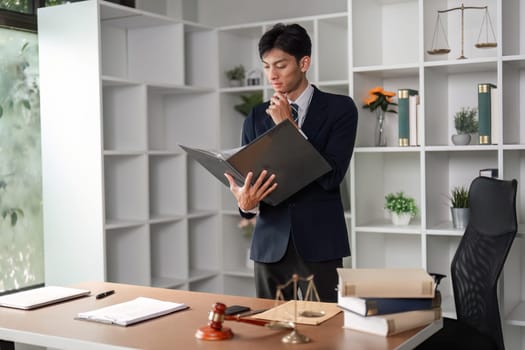 A thoughtful young Asian lawyer is analyzing case files and legal documents in a modern office setting, contemplating legal strategies.