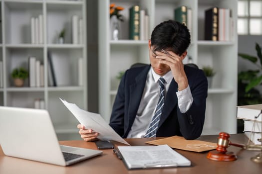 A young Asian lawyer appears stressed at his desk, reviewing legal documents and case files while managing challenges in his law practice.