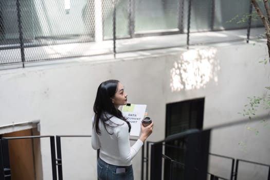 A young professional woman is holding a data report and a coffee cup, gazing upwards as she walks up the stairs in a contemporary office environment.