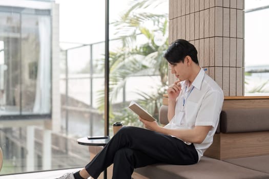 A young Asian male office worker sits in a contemporary workspace, thoughtfully reading a book and reflecting on personal development.