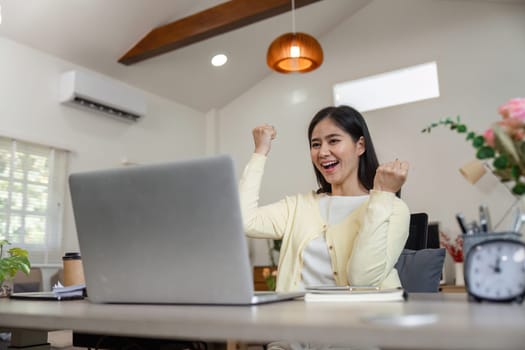 A joyful businesswoman celebrates her success while working on a laptop at home, embodying enthusiasm and achievement.