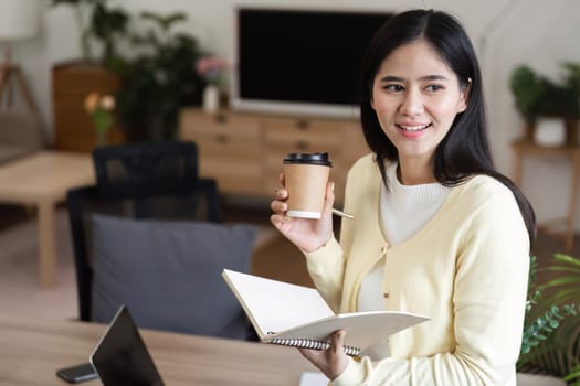 A businesswoman holds a coffee cup and notebook in her home office, preparing for a productive day.