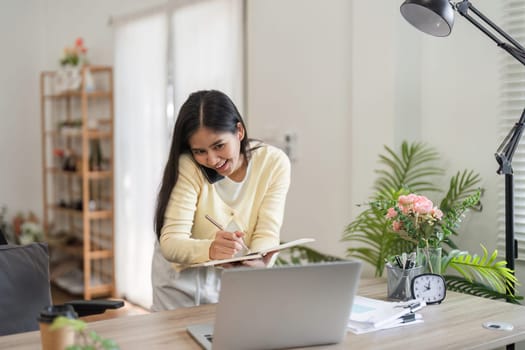 A focused young woman in a home office setting jots down notes while speaking on the phone, demonstrating effective multitasking skills.