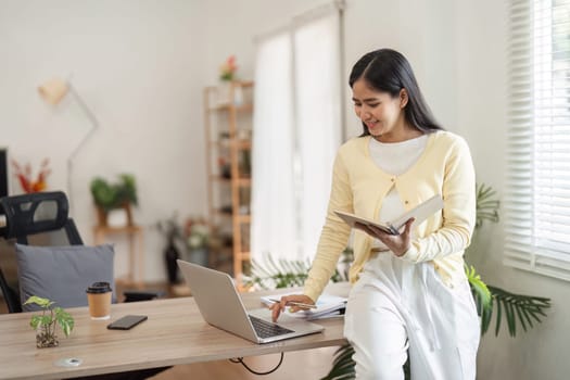 A young woman stands confidently by her desk, engaging with her laptop and notes, showcasing a productive home office setup.