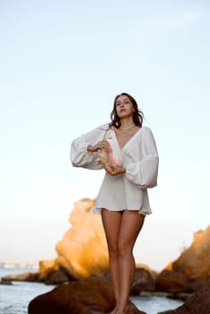 Young woman in summer white dress holds big shell in her hands. She is standing in sea, on beach. Behind her are rocks. Tourist trip abroad vacation, travel concept.