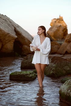 Young woman in a summer white dress walks along the beach near the sea in her hands she has a large shell, white with pink. she looks at the camera. Tourist trip abroad vacation, travel concept, sea vacation