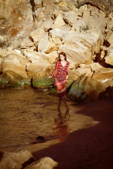 She is standing in the sea among huge rocks. Handmade dress, crocheted to fit. Boho style dress. She is walking along the seashore, general plan.