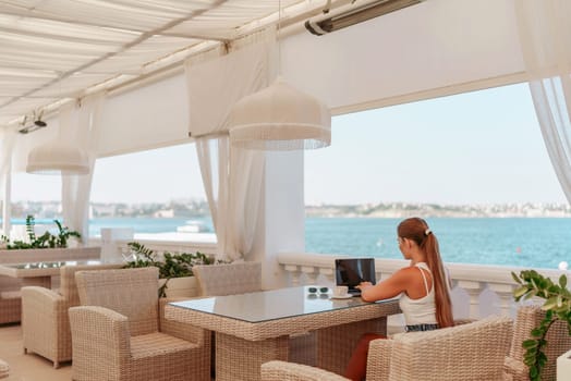 Woman coffee cafe laptop sea. Female freelancer using laptop on coffee break in modern cafe with sea view in the morning.
