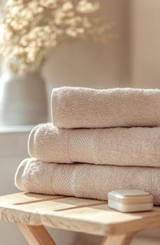 Three plush, beige towels are neatly stacked on a small wooden table near a bright window with soft sunlight filtering in.