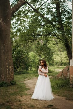 young girl bride in a white dress in a spring forest on a wedding day. beautiful brunette in a long stylish plan among huge trees.