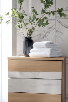 A clean bathroom setting features wooden storage, rolled white towels, and a decorative vase with green leaves for a fresh look.