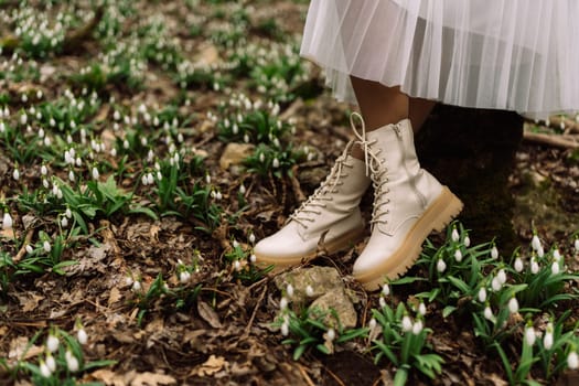 Cream-colored boots stand in a field of snowdrops. A woman's white skirt and legs are partially visible. Spring blooms carpet the forest floor.