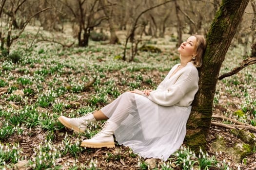 Woman in white dress sits against tree trunk in snowdrop-covered forest, eyes closed, basking in sunlight. Peaceful spring scene.