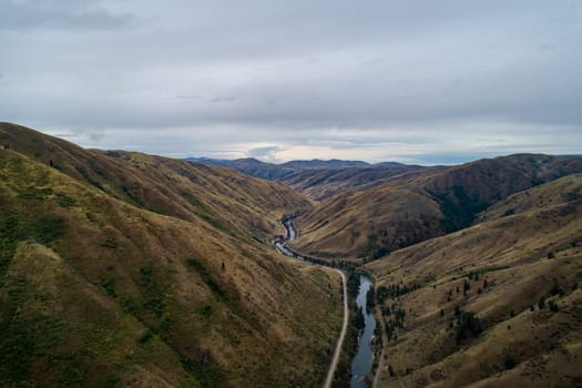 A mountain range with a river running through it. The sky is cloudy, and the landscape is mostly brown and green. Scene is peaceful and serene, with the river