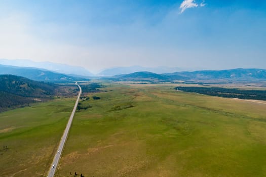 A road stretches across a vast, open field. The sky is clear and blue, and the mountains in the distance are covered in snow. The scene is peaceful and serene