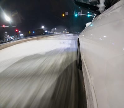 A car is driving down a snowy road at night. The car is white and has snow on the ground. The road is wet and slippery, and the car is driving at a slow speed. The scene is dark and quiet
