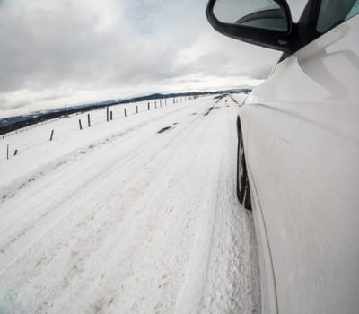A car is driving down a snowy road with a fence in the background. The car is in the driver's mirror and the snow is piled up on the road