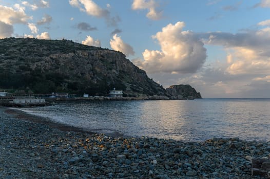 Gentle waves shimmer as twilight descends, reflecting clouds above a peaceful rocky beach beside a quiet village.