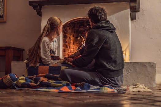 Family bonding by the fireplace during cozy winter evening. Warm and peaceful family moment indoors. Togetherness, love, and relaxation concept. A warm and inviting scene featuring a lit fireplace with glowing firewood and candles. A colorful traditional rug and pillow lie on the floor, creating a cozy atmosphere. Home, relaxation, and rustic comfort concept.