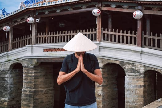 man traveler tourist in a Vietnamese hat prays at the ancient Japanese covered bridge in old town in Hoi An in Vietnam in Asia