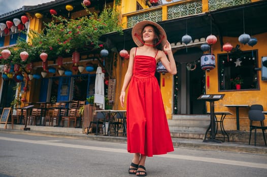 happy young female tourist traveler in traditional Vietnamese hat and red dress on street in old town in Hoi An city in Vietnam in Asia