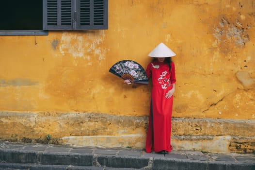 Asian young woman tourist in a traditional vietnamese hat and red dress with fan on old town street in Hoi An in Vietnam in Asia