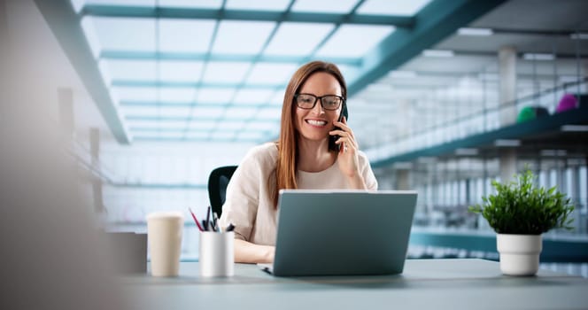 Busy woman managing phone and laptop in corporate office setting.
