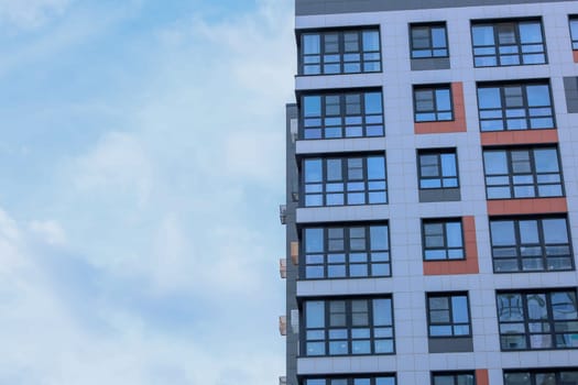 The concept of modern architecture, residential complex, apartment buildings for living. On the right, a close-up of a multi-storey, multi-apartment residential building in gray and white tones with orange inserts, large windows, panoramic balconies and baskets for air conditioning against the blue sky on the left with space for text. High quality photo