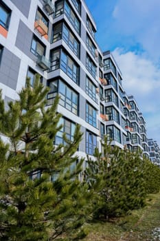 The concept of modern architecture, a residential complex, an apartment building for living. A multi-storey building in gray and white tones with orange inserts of modern architecture, with large windows and baskets for air conditioning, stands among coniferous trees against the background of a blue sky with clouds, view from below. High quality photo