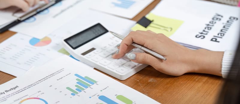 An office worker calculates financial data using a calculator, surrounded by charts and reports, highlighting budget planning efforts.