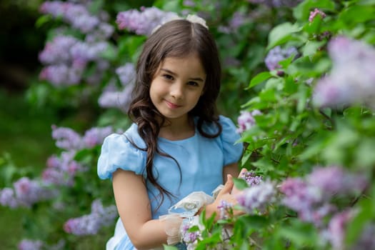 Young caucasian girl in blue dress smiling among lilac flowers in a garden.