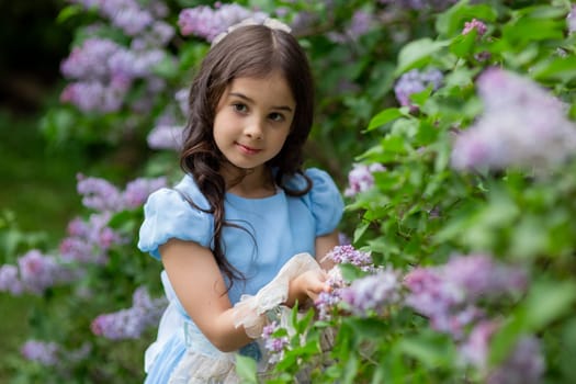 Young caucasian girl in blue dress among lilac blossoms in garden.