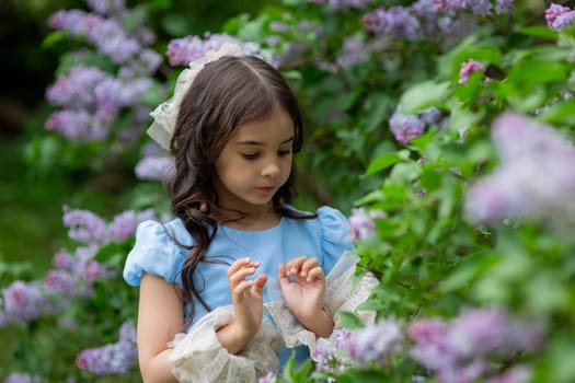 Caucasian young girl in blue dress surrounded by blooming lilac flowers in garden.